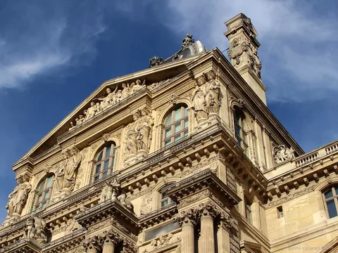 Fachada esculpida del Louvre con estatuas, frontón y columnas bajo cielo azul, vista con entradas Museo del Louvre