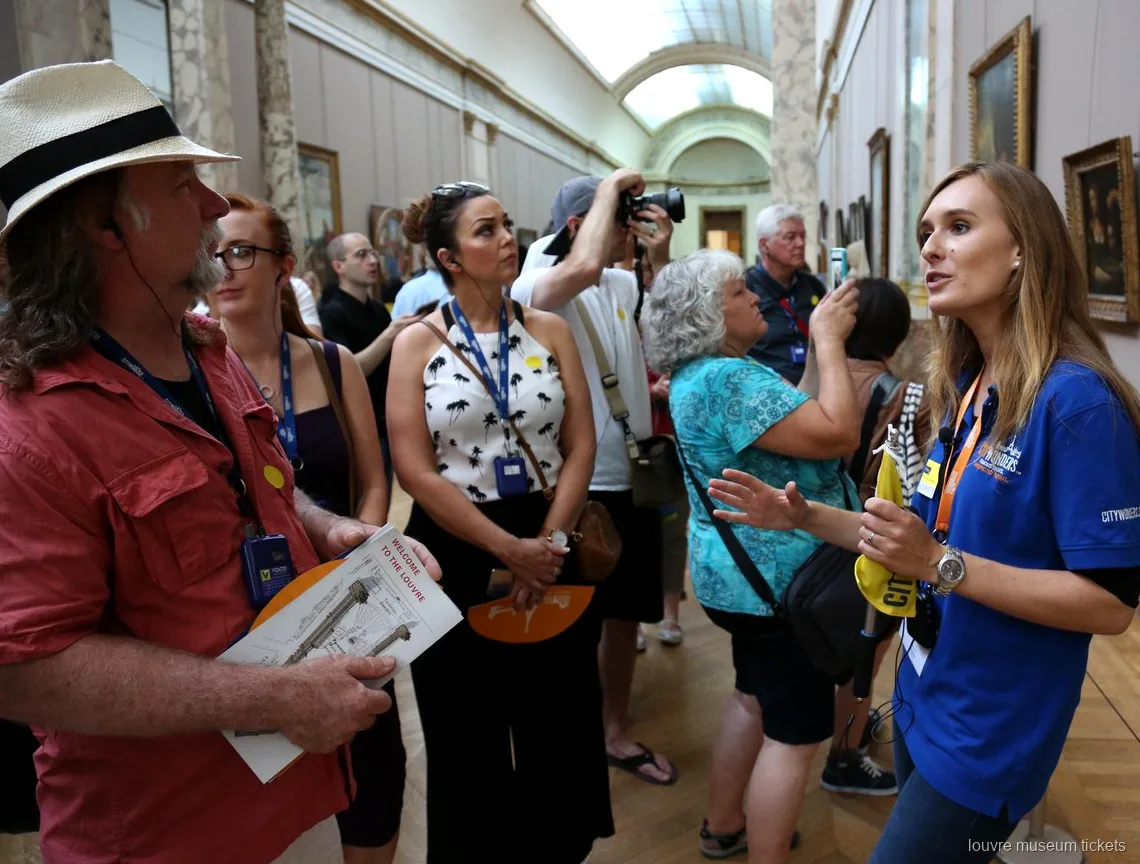 Guide in a blue shirt briefing a group holding Louvre maps in a painting gallery, tour with Louvre Museum tickets