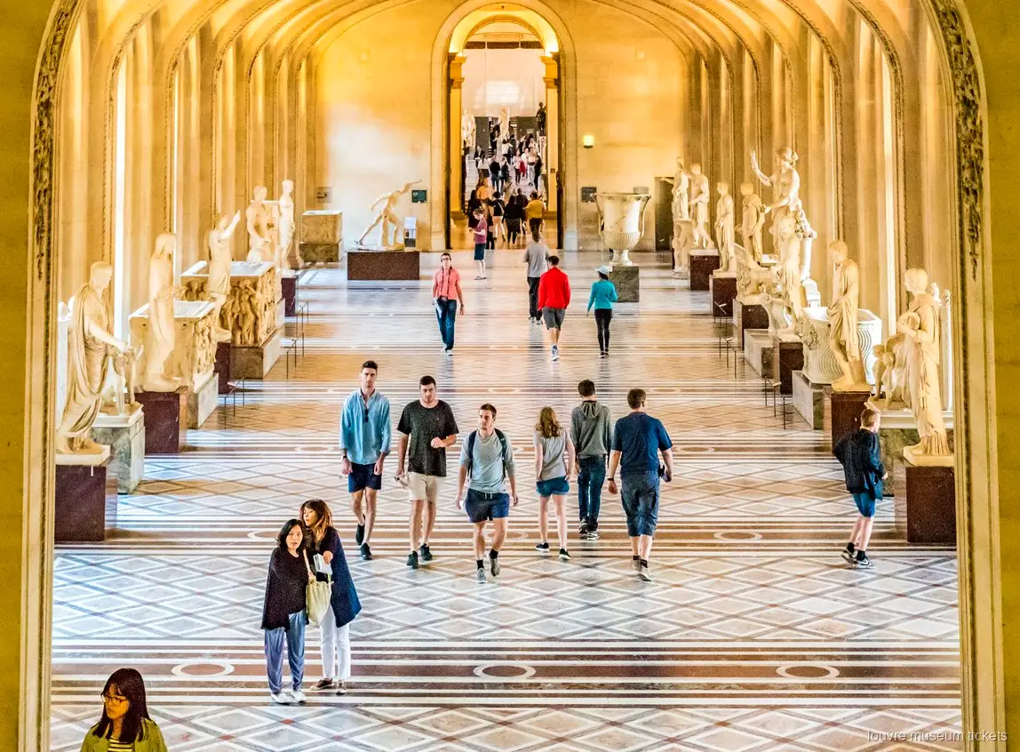 Visitors walking through a gallery of classical sculptures with a marble floor, seen with Louvre Museum tickets