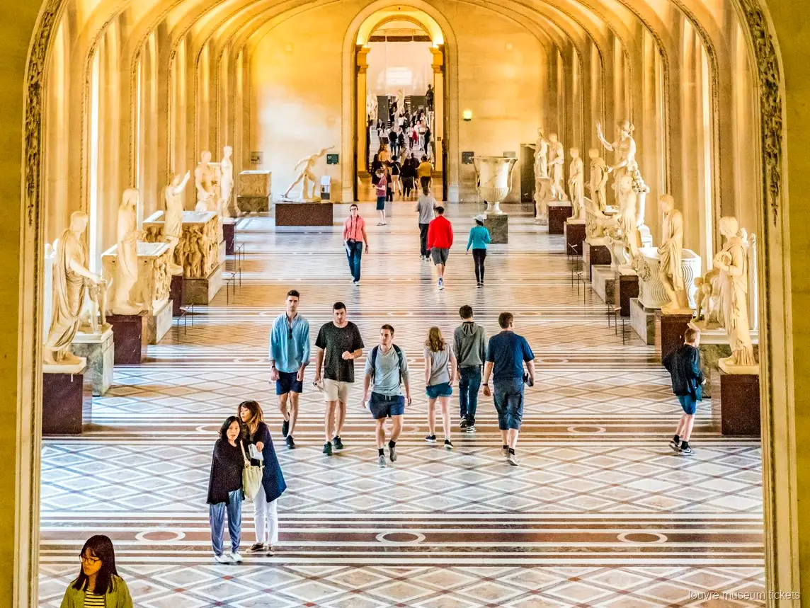 Kemerli ve mermer zeminli klasik heykel galerisinde yürüyen ziyaretçiler, Louvre Müzesi biletleri ile gezilen kanat