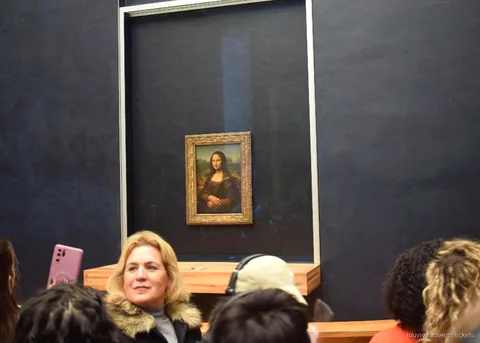 暗い壁のガラスケースに展示されたモナリザの前に集まる来館者、ルーブル美術館チケットで巡る小グループツアーの見どころ