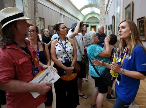 Guía con camiseta azul hablando a un grupo con planos del Louvre en una galería, tour con entradas Museo del Louvre